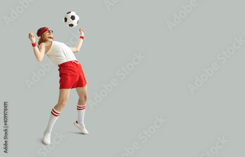 Fototapeta Naklejka Na Ścianę i Meble -  Funny nerdy man having fun with a football. Happy young male sports fan in white and red sportswear kicking away soccer ball on light gray background with free space for advertising text