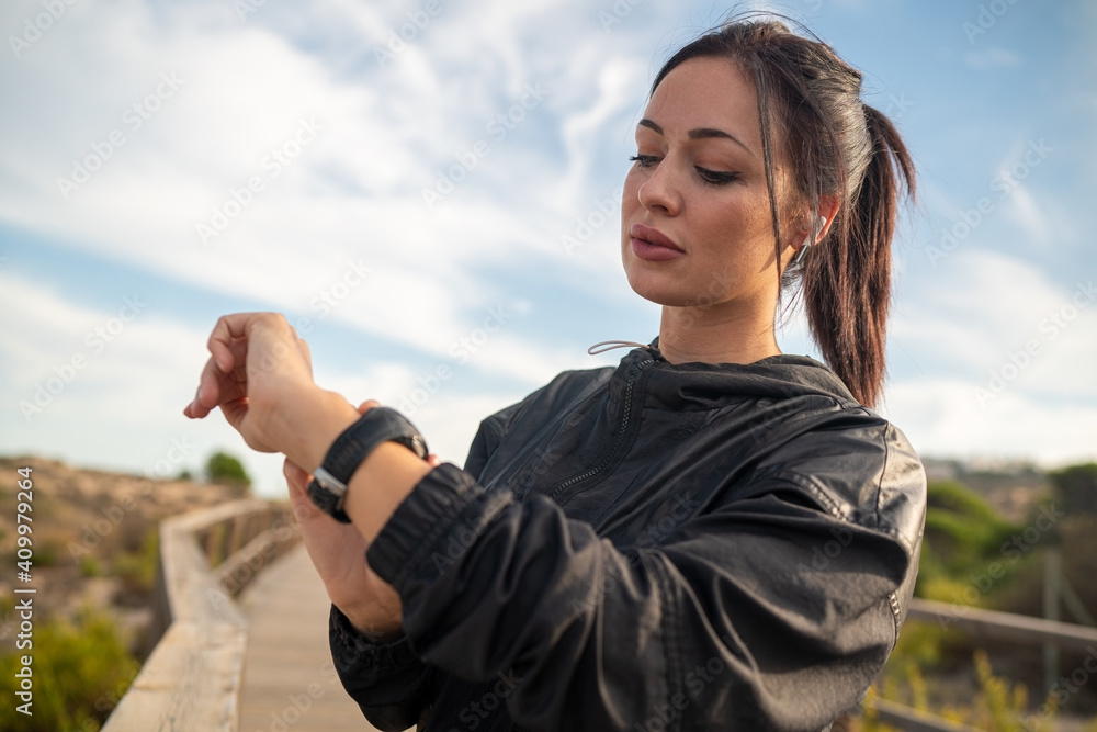 Active young brunette in black sports outfit standing on wooden pathway and checking data on fitness tracker after training in nature Active young brunette in black sports outfit standing on wooden pathway and checking data on fitness tracker after training in nature