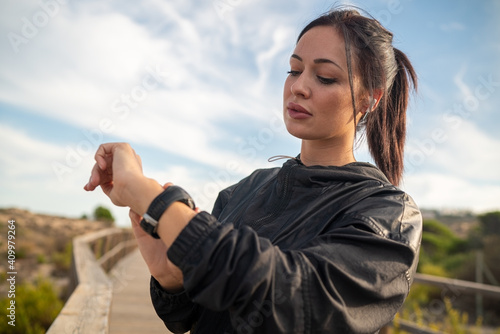 Active young brunette in black sports outfit standing on wooden pathway and checking data on fitness tracker after training in nature photo