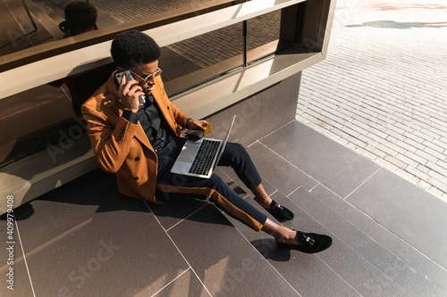 From above of concentrated young African American male freelancer in trendy suit and and sunglasses talking on smartphone and working on laptop while sitting on building stairs on street