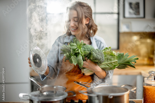 Pretty housewife in apron with fresh vegetables near cooker with boiling pans on the kitchen. Healthy green cooking concept. High quality photo