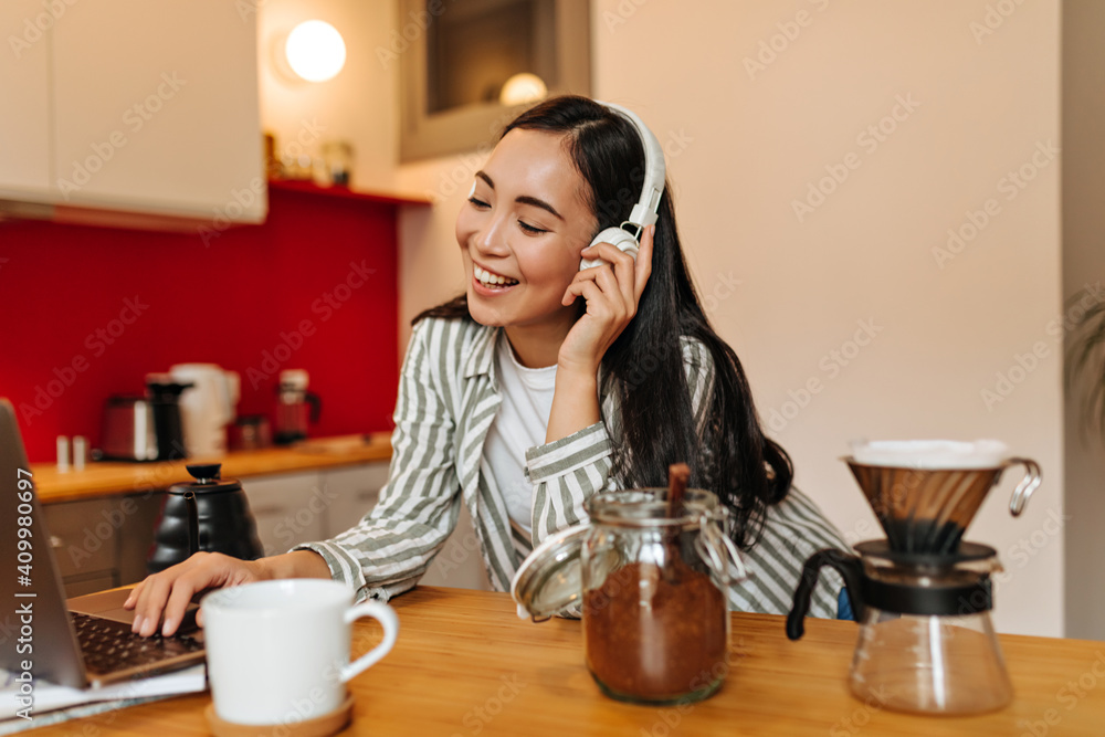 Beautiful Asian woman in white headphones smiling and looking into laptop screen Beautiful Asian woman in white headphones smiling and looking into laptop screen