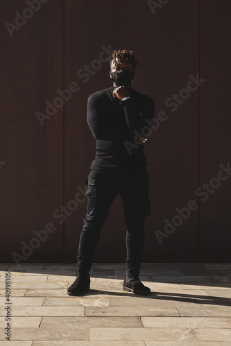 African American male in black outfit and protective mask standing on street and looking at camera during COVID 19 epidemic