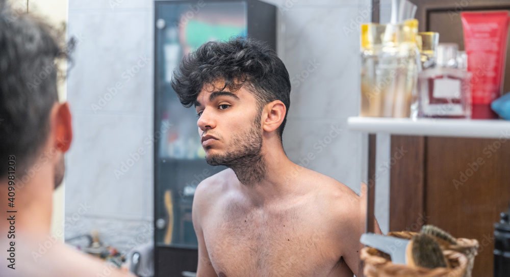 Shirtless young ethnic unshaven man looking at reflection in mirror during morning hygienic procedure in bathroom at home Shirtless young ethnic unshaven man looking at reflection in mirror during morning hygienic procedure in bathroom at home