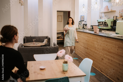 Positive female confectionery owner carrying plate with sweets while serving dessert for guest in cozy cafe