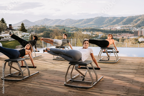 Company of people in sportswear doing abs exercises on pilates chair during training on terrace on sunny day photo