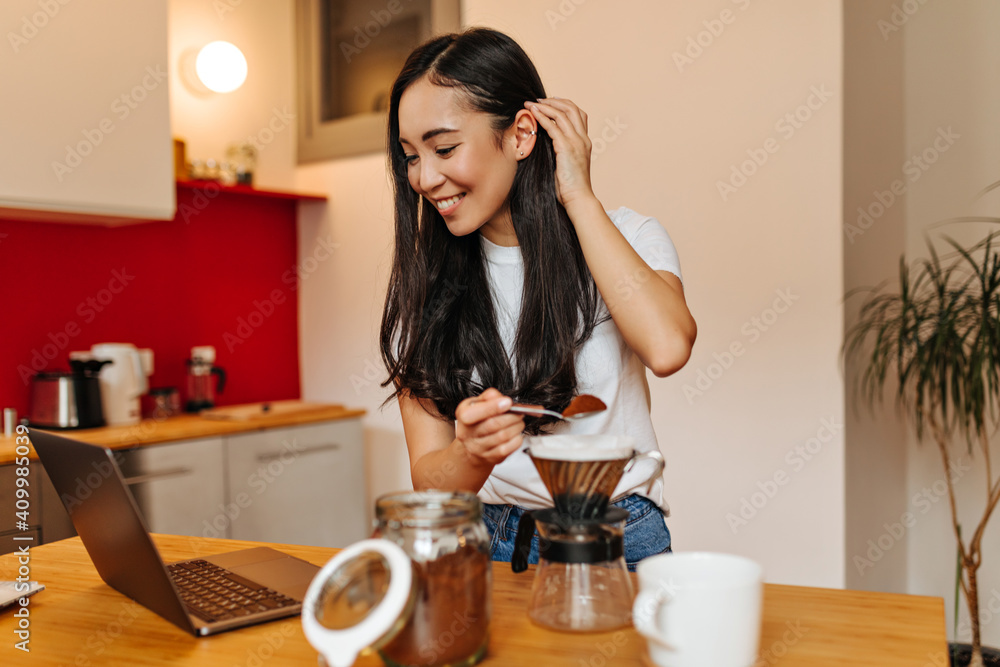 Joyful Asian girl makes natural coffee and watches video in laptop with smile Joyful Asian girl makes natural coffee and watches video in laptop with smile