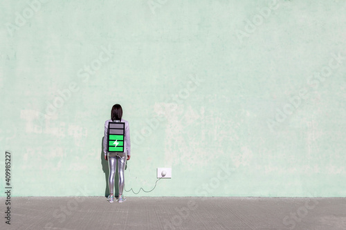 Back view of unrecognizable female with long dark hair in stylish outfit standing on street near wall with battery shaped backpack connected to charger