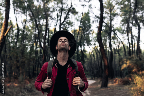 Adult traveler in hat closing eyes and breathing peacefully in harmony with nature of forest