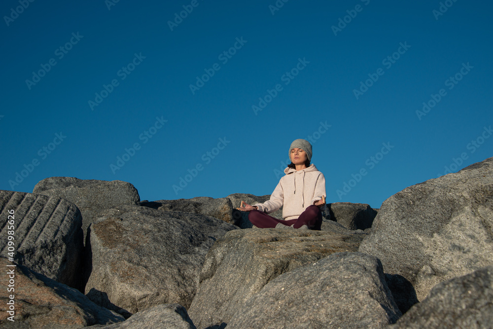 Young female in warm activewear and knitted hat meditating with mudra hand gesture while sitting on rock against blue sky Young female in warm activewear and knitted hat meditating with mudra hand gesture while sitting on rock against blue sky