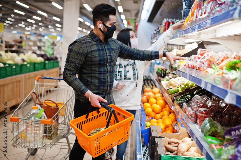 Asian couple wear in protective face mask shopping together in supermarket during pandemic. Taking vegetables from fridge.