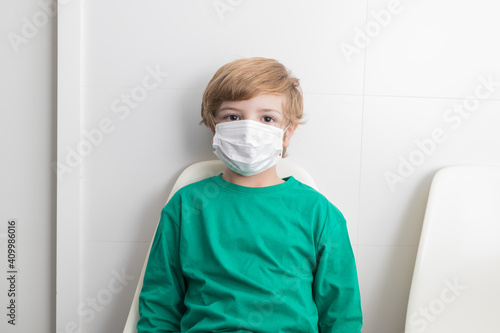 Calm boy in protective medical mask sitting on chair in medical room in clinic and looking at camera during COVID pandemic photo