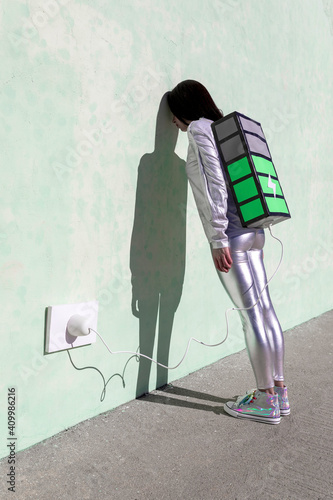 Side view of unrecognizable exhausted female in futuristic outfit leaning head on wall while standing on street and recharging energy