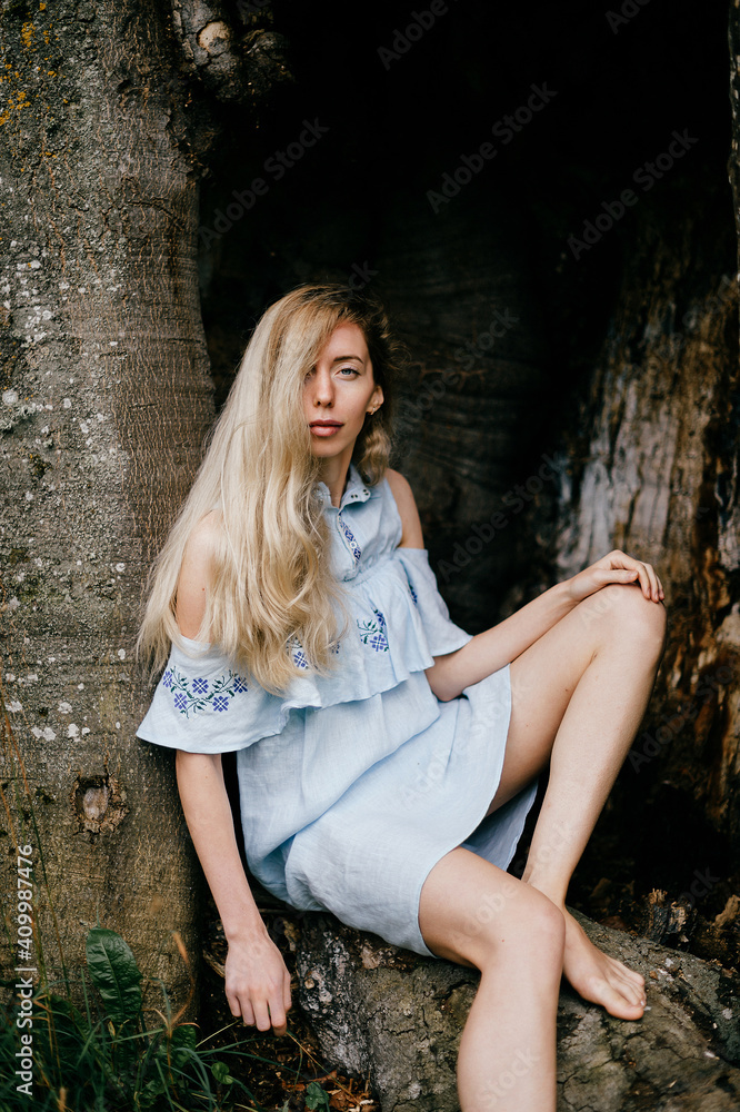 Fototapeta premium Young attractive barefoot blonde girl in blue romantic dress sitting in the old tree