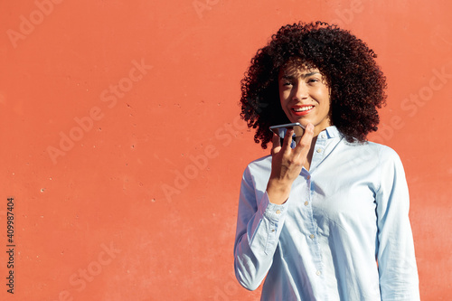 Content ethnic female entrepreneur in formal shirt recording voice message on smartphone while standing in city on orange background photo