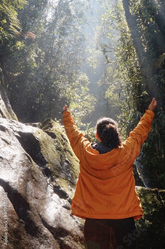 Brunette woman with curly hair and yellow jacket happy raising arms in the jungle