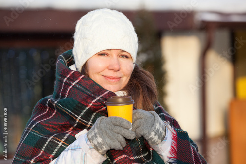 European woman in woollen gloves and checkered scarf holds a yellow paper cup of coffee on the winter city street. Hot and warming beverage in winter season © Kekyalyaynen