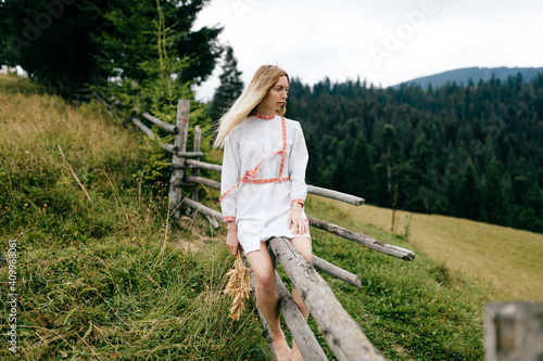 Young attractive blonde girl in white dress with ornament sitting on wooden fence with spikelets bouquet over picturesque countryside landscape