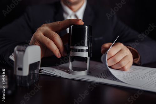 Businessman approves a profit contract. Stack of coins, contract and stamps on desk.