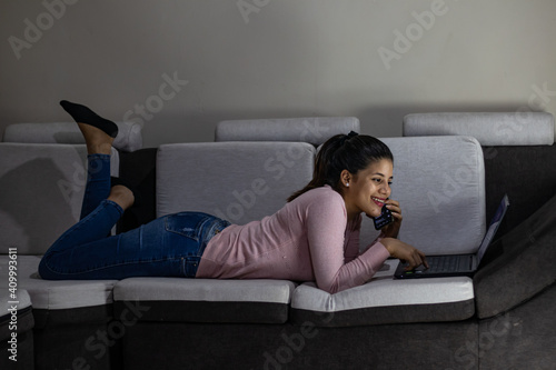 Smiling young woman lying on the sofa at home talking on cell phone