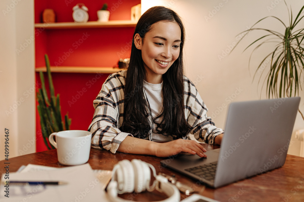 Fototapeta premium Brown-eyed woman in plaid jacket with smile working in laptop and posing at table with cup of coffee
