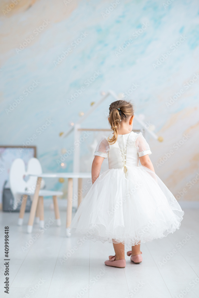 a little girl in a lush white dress and pink shoes stands with her back in a light room a little girl in a lush white dress and pink shoes stands with her back in a light room