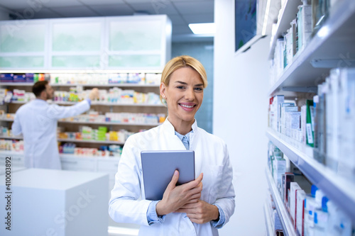 Portrait of beautiful female blonde pharmacist standing in pharmacy shop or drugstore by medicines and holding tablet. In background male druggist arranging boxes on the shelf.