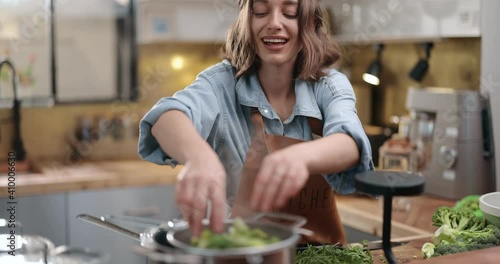 Young cheerful housewife in apron cooking broccoli on the kitchen at home. Happy household chores and healthy eating concept photo