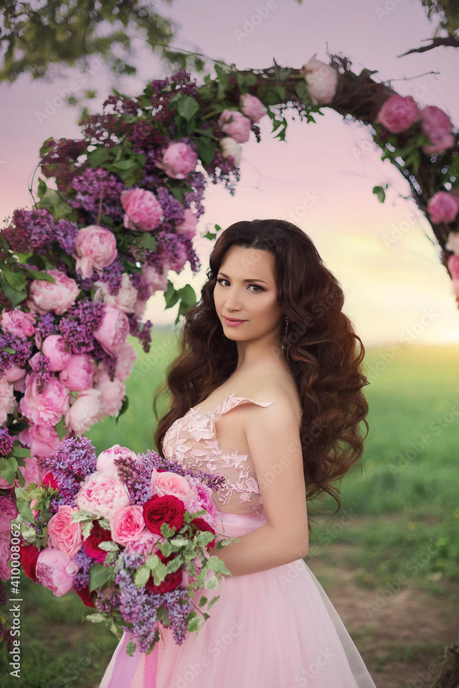 Fototapeta premium Beautiful bride in pink dress standing with her bouquet at a round flower arch under a large single tree in a field at sunset. A destination summer wedding, romantic country ceremony.