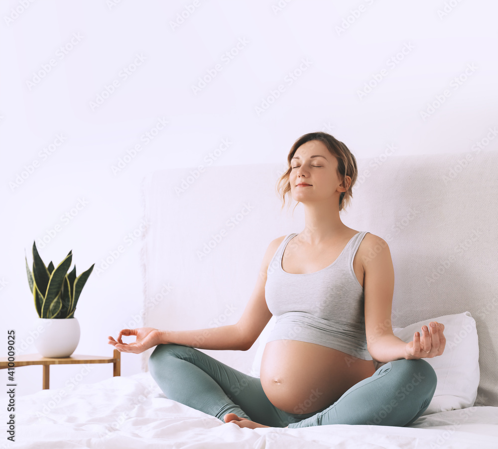 Pregnant woman in lotus pose doing meditation or breathing exercises for healthy pregnancy and preparing body for childbirth. Young expectant mother practicing yoga at home. Pregnant woman in lotus pose doing meditation or breathing exercises for healthy pregnancy and preparing body for childbirth. Young expectant mother practicing yoga at home.