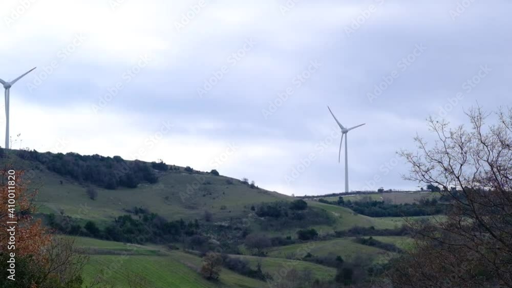 Slow motion video. Wind turbine towers top of the small hills and agricultural field before harvesting in very cloudy sky background. Slow motion video. Wind turbine towers top of the small hills and agricultural field before harvesting in very cloudy sky background.