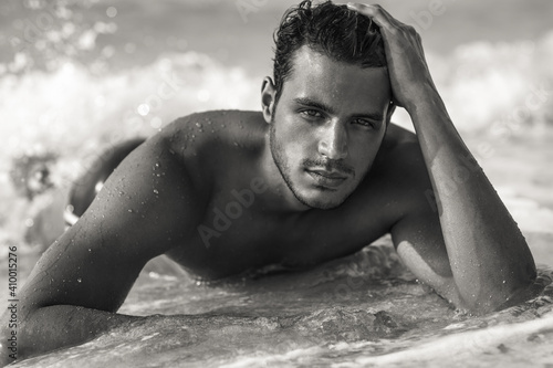 Portrait of sexy handsome topless male model with beautiful eyes laying in the ocean water on the beach. Black and white.
