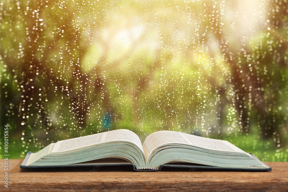 The open book which there is on a table on window glass with raindrops after summer rain - obrazy, fototapety, plakaty 
