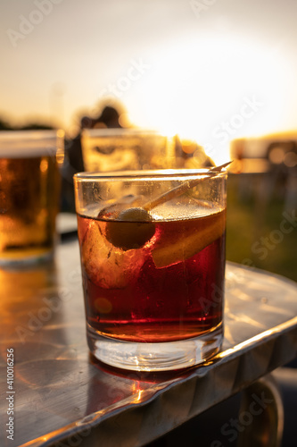 glass of vermouth with ice and a green olive inside on a steel table with people and sun on the background