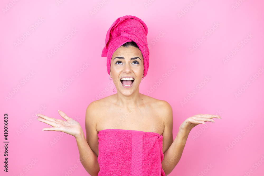 Young beautiful woman wearing shower towel after bath standing over isolated pink background clueless and confused expression with arms and hands raised Young beautiful woman wearing shower towel after bath standing over isolated pink background clueless and confused expression with arms and hands raised