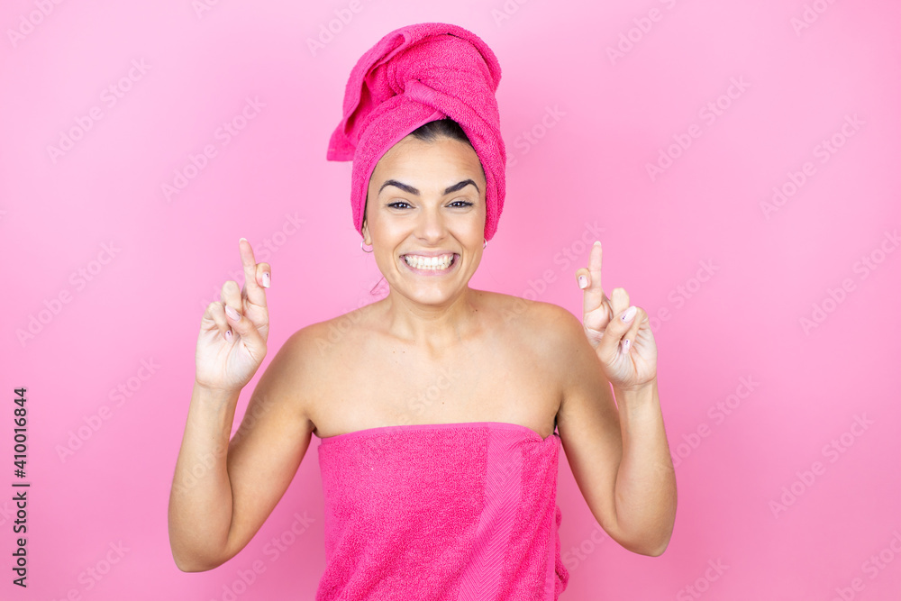 Young beautiful woman wearing shower towel after bath standing over isolated pink background gesturing finger crossed smiling with hope and looking side Young beautiful woman wearing shower towel after bath standing over isolated pink background gesturing finger crossed smiling with hope and looking side