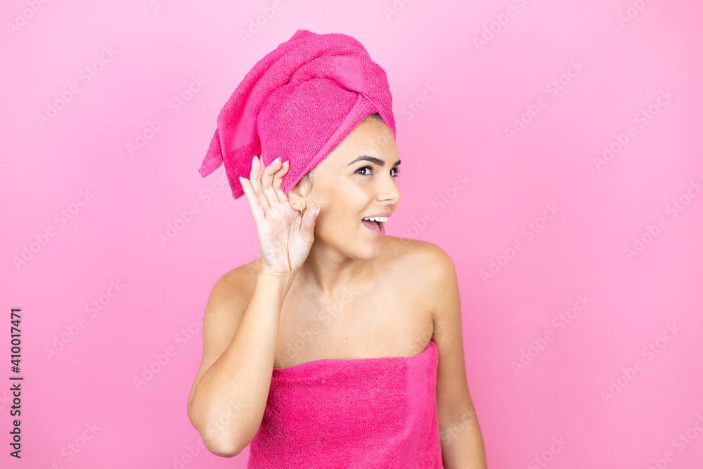 Young beautiful woman wearing shower towel after bath standing over isolated pink background surprised with hand over ear listening an hearing to rumor or gossip Young beautiful woman wearing shower towel after bath standing over isolated pink background surprised with hand over ear listening an hearing to rumor or gossip