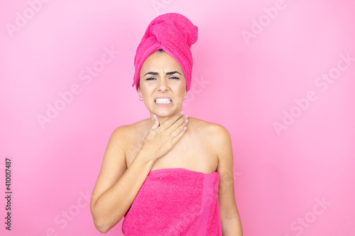 Young beautiful woman wearing shower towel after bath standing over isolated pink background touching painful neck, sore throat for flu, clod and infection