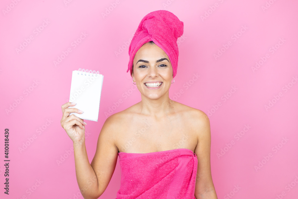 Young beautiful woman wearing shower towel after bath standing over isolated pink background smiling and showing blank notebook in her hand Young beautiful woman wearing shower towel after bath standing over isolated pink background smiling and showing blank notebook in her hand