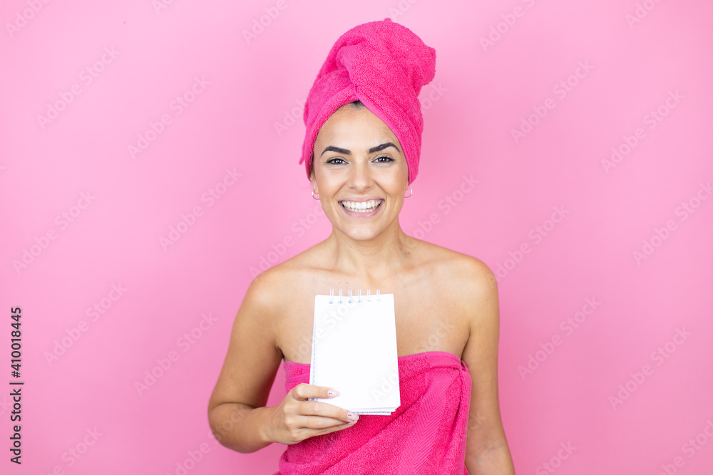 Young beautiful woman wearing shower towel after bath standing over isolated pink background smiling and showing blank notebook in her hand Young beautiful woman wearing shower towel after bath standing over isolated pink background smiling and showing blank notebook in her hand