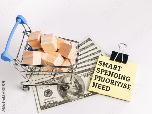 Business and shopping concept. Selective focus a bunch of wooden cubes in trolley with fake money and text SMART SPENDING PRIORITIZE NEED on sticky note.