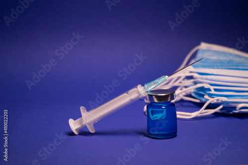 Vaccine bottle phial with no label and medical syringe with injection needle near pile of blue medical masks . isolated on blue background. cure. Development of coronavirus vaccine COVID-19