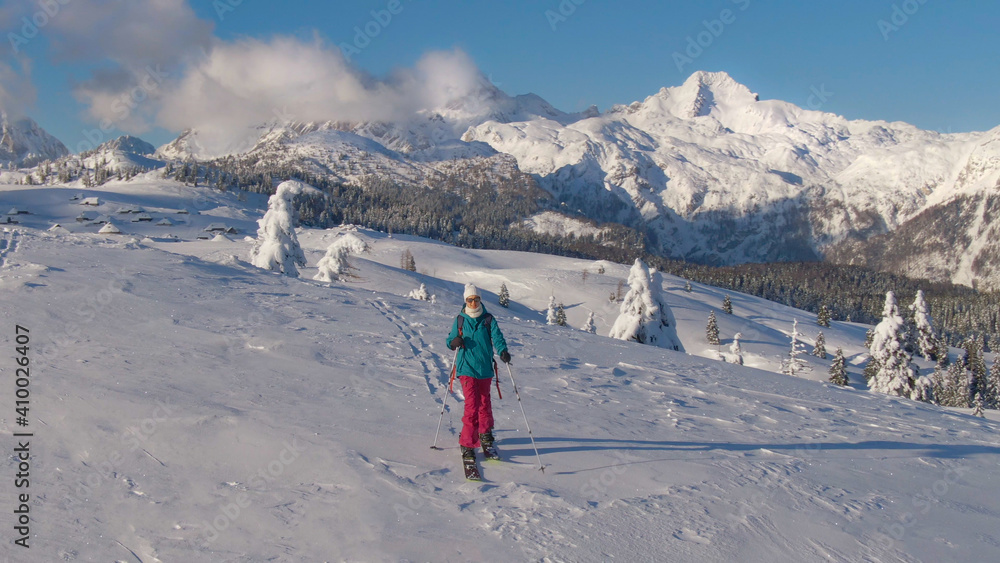 AERIAL: Female ski tourer hikes across a snowy meadow in wintry Velika Planina.
