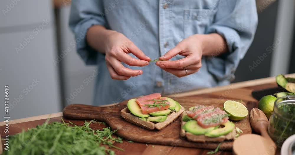 Woman making healthy snacks made of avocado and salmon on the kitchen with green ingredients on the table. Putting greens on toasts. Close-up shot Woman making healthy snacks made of avocado and salmon on the kitchen with green ingredients on the table. Putting greens on toasts. Close-up shot