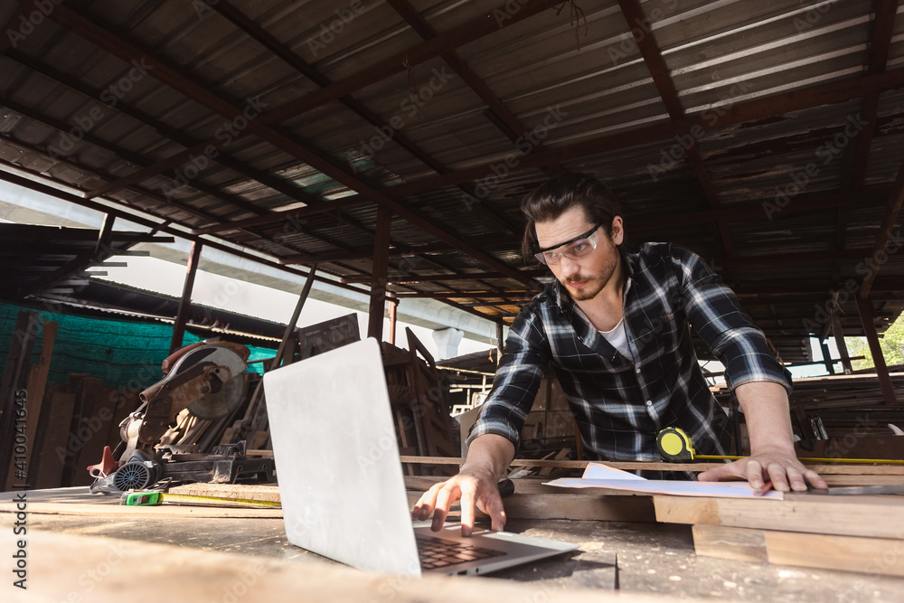 young carpenter caucasoid man worker. he looking designs in laptop computer at own workspace. craftsman profession in wood factory. young carpenter caucasoid man worker. he looking designs in laptop computer at own workspace. craftsman profession in wood factory.