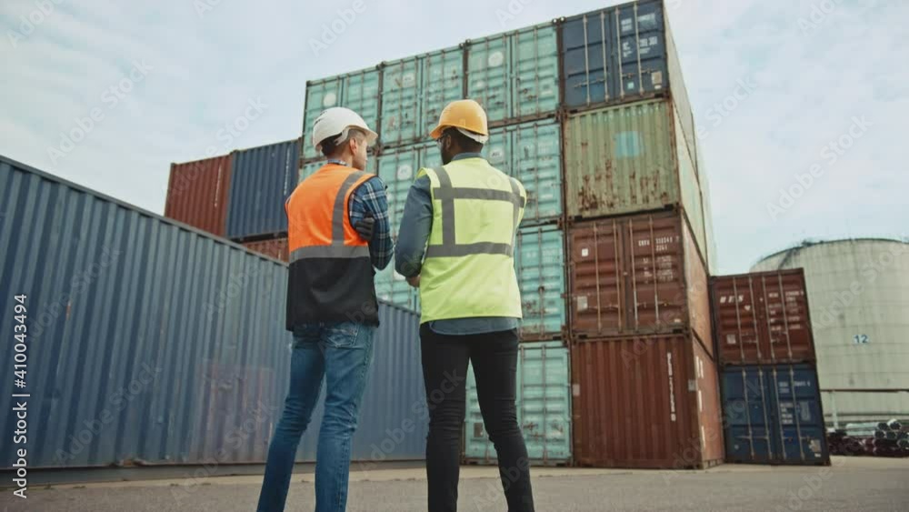 Handsome Industrial Engineer and African American Supervisor in Hard Hats and Safety Vests Standing with Their Backs to the Camera in Container Terminal. Colleagues Talk About Logistics Operations. Handsome Industrial Engineer and African American Supervisor in Hard Hats and Safety Vests Standing with Their Backs to the Camera in Container Terminal. Colleagues Talk About Logistics Operations.