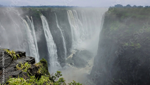 Powerful streams of water rush down from the rocks. In a deep gorge  there is fog from water splashes. In the foreground  wet stones and green grass. Victoria Falls. Zimbabwe.