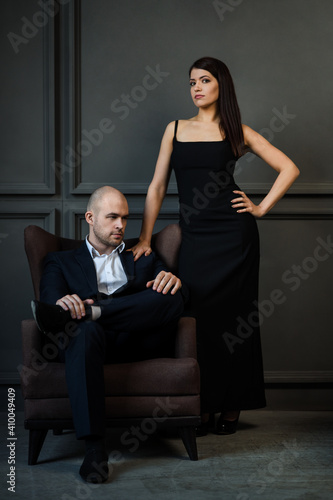 Elegant couple in love. A man and a woman in a dark room. The girl in the black dress is standing and the bald guy in a suit sitting in a chair, loft. Vertical photo. Valentine's Day