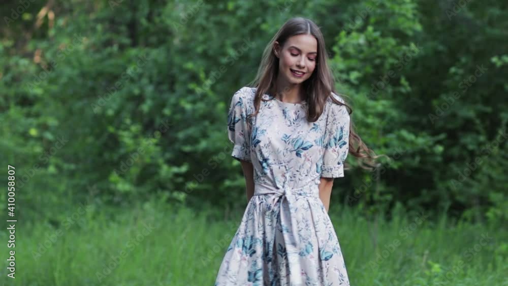 A beautiful woman smiles as she walks along a path surrounded by foliage and trees. Front view of a woman in a dress walking in a summer Park. Slow motion A beautiful woman smiles as she walks along a path surrounded by foliage and trees. Front view of a woman in a dress walking in a summer Park. Slow motion