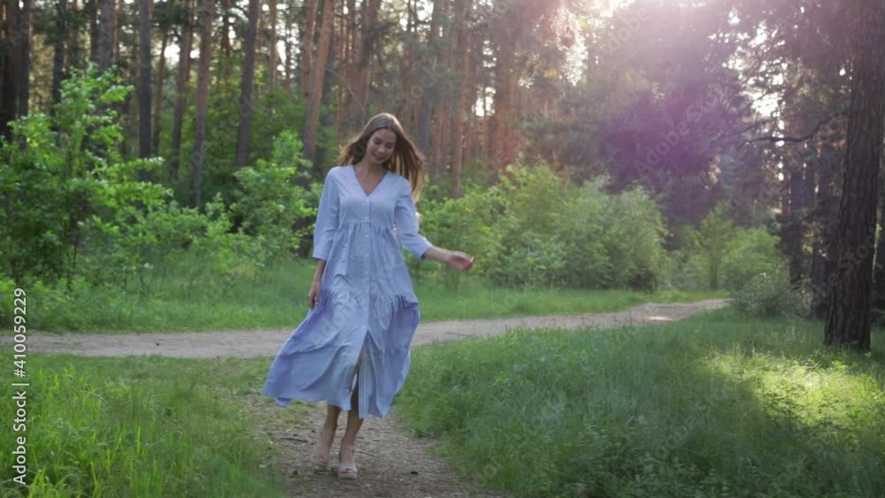 A beautiful woman smiles as she walks along a path surrounded by foliage and trees. Front view of a woman in a dress walking in a summer Park. Slow motion A beautiful woman smiles as she walks along a path surrounded by foliage and trees. Front view of a woman in a dress walking in a summer Park. Slow motion